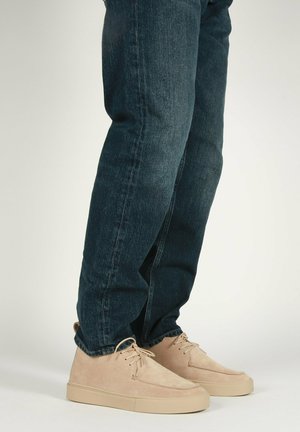 Sneaker in suede beige con un design a profilo basso, con punta arrotondata e suola in gomma beige, abbinate a jeans denim blu scuro.