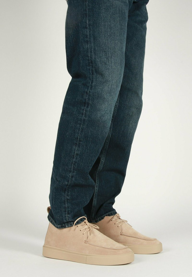 Sneaker in suede beige con un design a profilo basso, con punta arrotondata e suola in gomma beige, abbinate a jeans denim blu scuro.