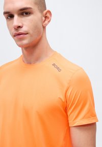 Νεαρός άνδρας με ανοιχτόχρωμο δέρμα και κοντά μαλλιά, φοράει πορτοκαλί κοντομάνικο πουκάμισο με το λογότυπο Borg, κοιτάζοντας ελαφρώς πλάγια.