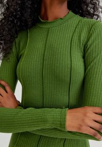 Haut vert à manches longues côtelé, à la coupe ajustée, avec un col montant et des détails de coutures. Texture douce avec une légère élasticité.