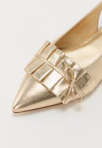 Chaussure dorée à bout pointu avec une semelle plate, présentant une sangle plissée à l'avant et un petit nœud. Fini métallique, texture lisse.