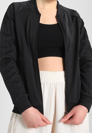 Czarna sportowa kurtka z textured mesh rękawami, zamek z przodu i żeberkowymi mankietami. Połączona z czarnym topem krótkim i jasnymi spodniami.