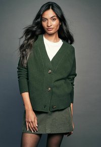 Grön stickad cardigan med ribbade detaljer, med tre knäpptillverkade knappar. Kombinerad med en vit skjorta och en grön kordskärning. Enkelt design.