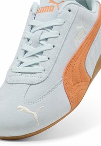 Tenisici din suede albastru deschis cu accente portocalii, prezentând detalii albe ale logo-ului, sireturi plate și o talpă din cauciuc de tip gumă.