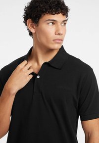 Polo shirt nera realizzata in tessuto morbido, con colletto, maniche corte, chiusura a bottoni e logo "GUESS JEANS" in rilievo sul petto.