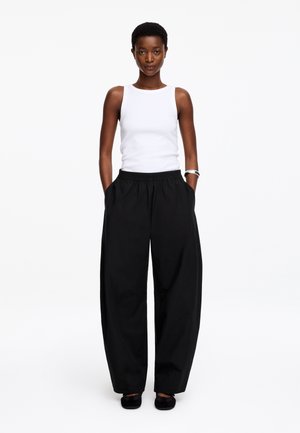 Femme debout, mains dans les poches, portant un haut blanc sans manches, un pantalon large noir, des chaussures noires et un bracelet argenté au poignet gauche.