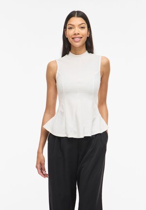 Femme souriante portant un top peplum blanc sans manches et un pantalon noir ample, debout devant un fond blanc uni.
