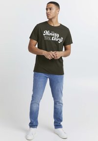 Oliven grønn bomulls-T-skjorte med hvit grafisk tekst som leser "EST. Ninety th3r3 Blend Denim." Kombinert med lyseblå jeans og hvite joggesko.