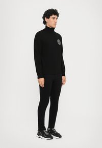Μαύρος πουλόβερ με ψηλό λαιμό και στρογγυλό λογότυπο στο στήθος, συνδυασμένος με μαύρο παντελόνι slim-fit και μαύρα αθλητικά παπούτσια με άσπρες λεπτομέρειες.