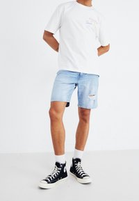 Camiseta blanca de manga corta con texto multicolor; pantalones cortos de mezclilla azul claro con dobladillo deshilachado; zapatillas negras de caña alta con detalles blancos.