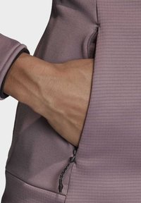 Fialová sportovní mikina s ribkovanou texturou, která má boční kapsu a ruku vloženou do kapsy, při�čemž ukazuje detail zipu kapsy.