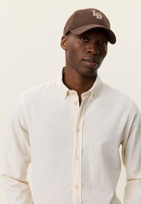 Καφέ μπέιζμπολ καπέλο με λογότυπο, συνδυασμένο με ανοιχτό μπεζ πουκάμισο με κουμπιά. Το πουκάμισο έχει απαλό υφή και διακριτικό ανάγλυφο σχέδιο.