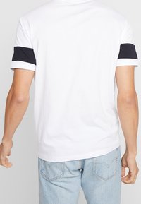 Bílá tričko s černým horizontálním pruhem na rukávech, měkká látka, kulatý výstřih, volný střih, v kombinaci s světle modrými džíny.