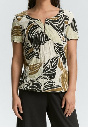 Kvinde iført kortærmet bluse med sort, brun og hvid abstrakt bladmønster og små knapper foran, kombineret med sorte bukser.