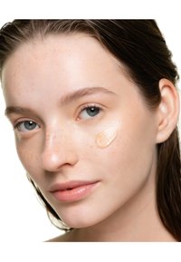 Visage avec de légères taches de rousseur, peau lisse et éclat naturel, affichant une trace de maquillage beige sur la joue. De longs cheveux foncés sont partiellement visibles.