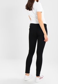 Žena oblečená v černých skinny džínách, bílém tričku s krátkým rukávem a bílých teniskách s červenými květinovými vzory, stojící na jednobarevném světlém pozadí.