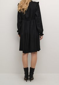 Svart satin klänning med figursydd midja, långa ärmar och volangdetaljer på axlarna, kombinerad med svarta klackstövlar.