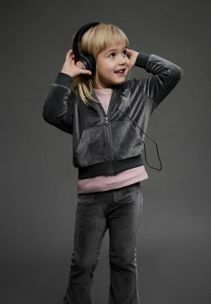 Grå velour træningsdragt med zip-jakke og matchende bukser. Barnet har sorte headphones på, smiler; lyserød skjorte under.