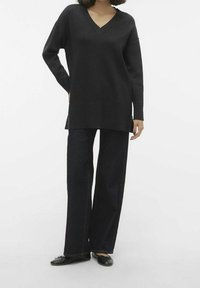 Maglione oversized nero a V con maniche lunghe, abbinato a jeans scuri a gamba larga e ballerine nere su uno sfondo semplice.