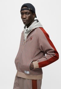 Φούτερ με φερμουάρ και σχέδιο houndstooth σε κόκκινο, γκρι και μπεζ, με γκρι κουκούλα, ριμπ μανσέτες και υφασμάτινη ετικέτα. Τοποθετημένο σε ουδέτερο φόντο.