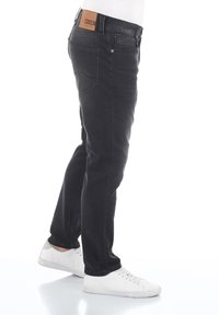 Jeans de mezclilla negra con un ajuste slim, que cuentan con una etiqueta de cuero en la cintura, diseño de cinco bolsillos y detalles de costura mínimos. Combinados con zapatillas blancas.