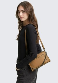 Tan leren crossbody tas met een gestructureerde afwerking, voorzien van metalen kettingaccenten en een rechthoekige vorm; gedragen op een zwarte top en donkere jeans.