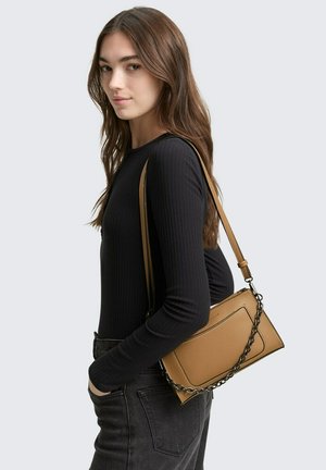Beige Leder-Crossbody-Tasche mit strukturiertem Finish, ausgestattet mit metallischen Kettenakzenten und rechteckiger Form; getragen zu einem schwarzen Oberteil und dunklen Jeans.