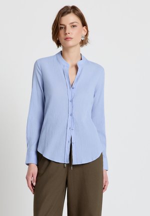 Woman wearing light blue long-sleeve textured blouse with mandarin collar and olive green wide-leg pants, standing against plain background.