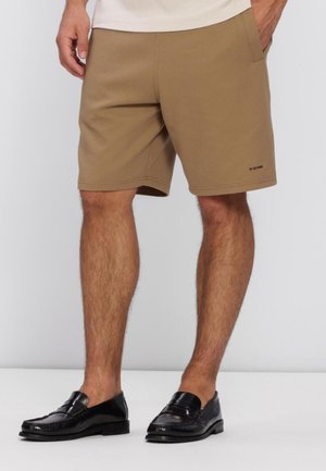 Uomo con pantaloncini color nocciola dalla texture strutturata e mocassini neri in pelle, in piedi con una mano in tasca su sfondo semplice.