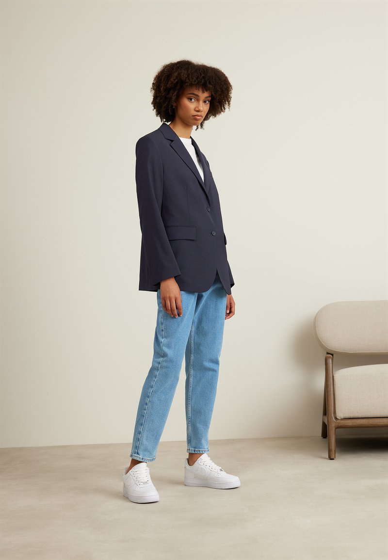 Blazer marine ajusté avec deux boutons, associé à un jean droit bleu clair et des baskets blanches. Éléments de design simples et modernes.