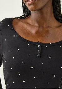 Sort langærmet skjorte med sølvstjerne mønstre, scoop neckline og tre sorte knapper foran. Blødt, strækbart stof.