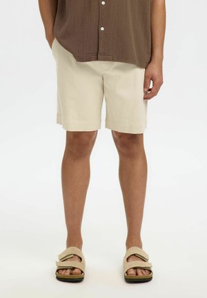 Homme portant un short beige, une chemise marron boutonnée et des sandales beige clair, debout devant un fond blanc.