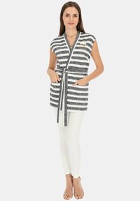Czarna i biała, w paski, bezrękawnikowa bluzka typu wrap z paskiem do wiązania, z kieszeniami i teksturowanym wykończeniem. Stylizowana z białymi spodniami.