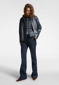 Giacca puffer blu navy con cappuccio, chiusura con zip e tasche laterali, indossata sopra una camicia a quadri. Pantaloni in denim scuri svasati con scarpe a punta.