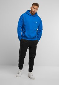 Sudadera con capucha azul y bolsillo frontal, combinada con joggers negros y zapatillas blancas. Diseño simple, tejido suave.