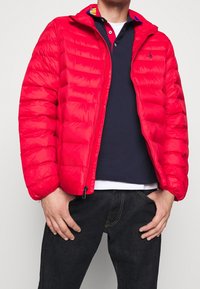 Röd puffjacka med horisontell quiltning, dragkedja och huva. Bärs över en marinblå polo med kontrasterande krage och vit nederkant.