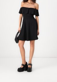 Černé šaty bez ramínek s volánkovým výstřihem, přiléhavým korzetem a rozšířenou sukní. V páru s černými platformovými botami se sponou.