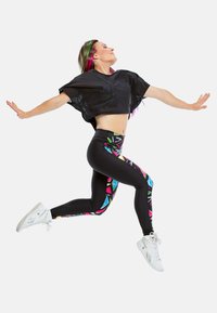Czarny crop top z luźnymi rękawami, w połączeniu z dopasowanymi czarnymi legginsami z kolorowymi geometrycznymi wzorami. Białe buty sportowe.