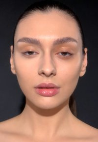 Close-up portrait with fair skin, defined eyebrows, and glossy nude lips. Background is dark, highlighting facial features and smooth texture.