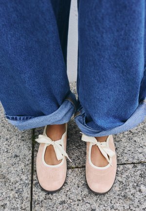 Nohy v světle růžových baletních botách se smetanovými mašlemi, ve světle modrých zavinutých džínách, stojící na šedých dlaždicích na chodníku.