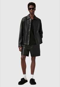 Black leather jacket with a collared neckline, button front, and matching leather shorts. Model wears dark sunglasses and black footwear.