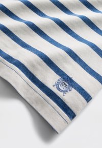 Bomullsstoff med horisontale marineblå og kremhvite striper, med en brodert emblem i nederste hjørne. Glatt tekstur.