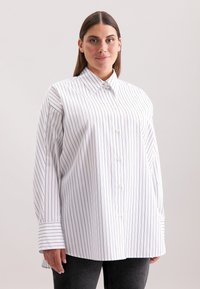 Camicia bianca con righe verticali blu navy, dotata di colletto appuntito e maniche lunghe con polsini larghi. Tessuto morbido con una superficie liscia.