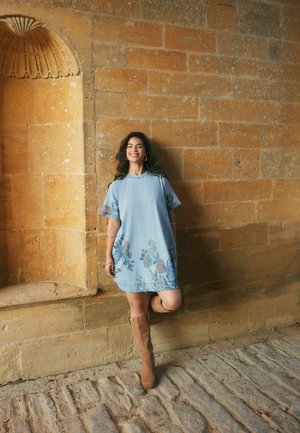 Noor naine sinise lillemustrilise kleidi ja beežide põlvikutega toetub struktuurse kiviseina vastu, naeratades ühe põlvega kõverdatud munakivisillutisel.