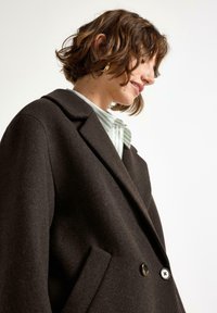 Cappotto di lana marrone scuro con una vestibilità strutturata, grandi rever oversize e due bottoni frontali di grandi dimensioni; presenta tasche laterali e una texture morbida.