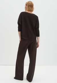 Frau mit kurzen blonden Haaren, die ein lockeres, dunkelbraunes Oberteil und weite Hosen trägt, steht mit dem Rücken zu einem schlichten, hellen Hintergrund.