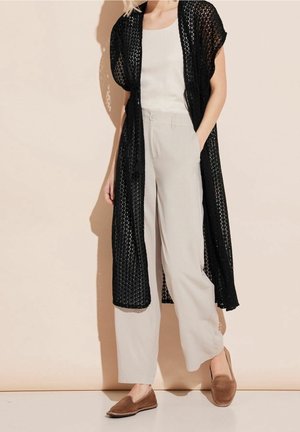 Vrouw draagt beige wijde broek, wit mouwloos topje, lange zwarte opengewerkte cardigan en bruine instapschoenen tegen een beige achtergrond.