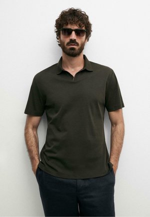 Mann mit lockigem Haar und Bart, der ein dunkelgrünes Polohemd, schwarze Sonnenbrille und dunkle Hose trägt, steht vor einer weißen Wand.