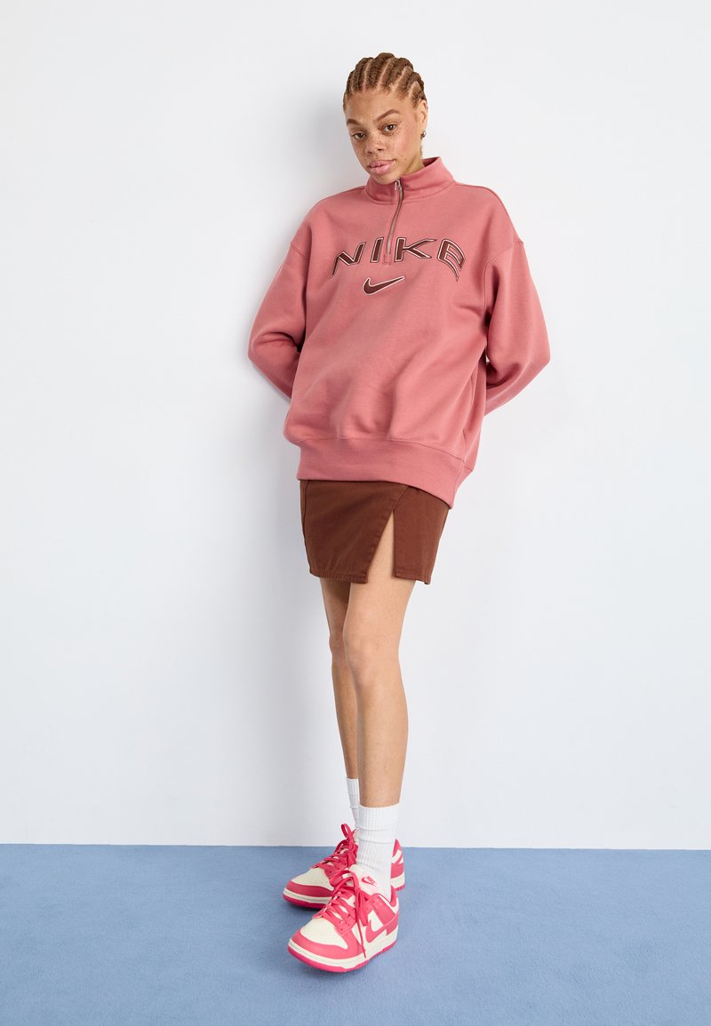 Helle rosa Nike-Pulloverjacke mit Reißverschluss, brauer Minirock mit seitlichem Schlitz und pinke Nike-Schuhe kombiniert mit weißen Socken vor einem blauen Hintergrund.
