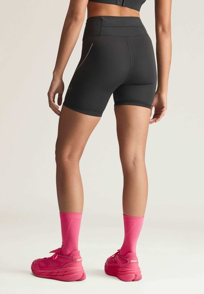 Woman wearing black high-waisted athletic shorts, pink crew socks, and pink running shoes, standing facing away on a light background.